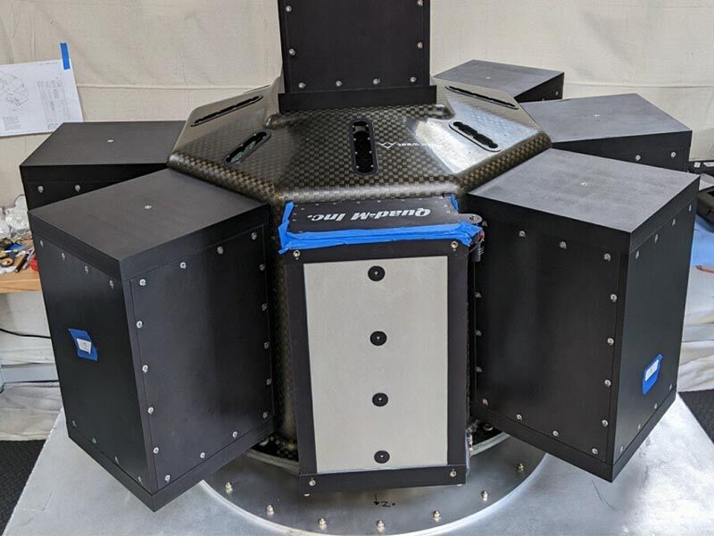 Image is in a manufacturing area. The launch adapter structure is sitting on a table top. Several surfaces of the octagonal structure have cube-shaped smallsats attached. Six smallsats are visible on the sides of the structure and one  is attached to the top. (Image courtesy QuadM)